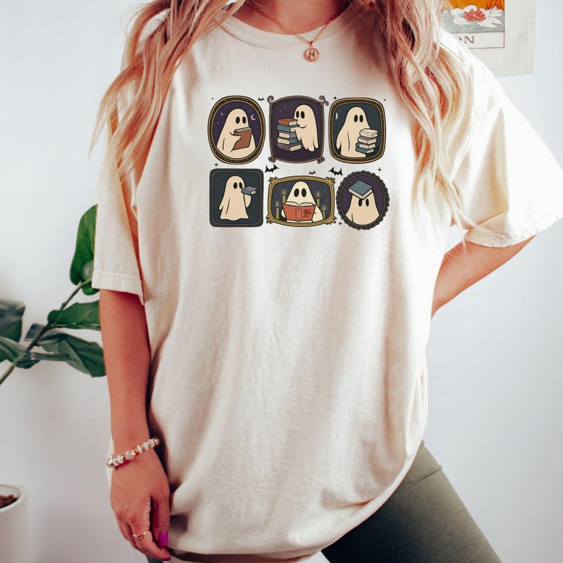 May include: A cream-coloured t-shirt with a graphic of six cartoon ghosts in frames. Each ghost is shown reading books or holding a book. The frames are in various shapes, with a vintage aesthetic.