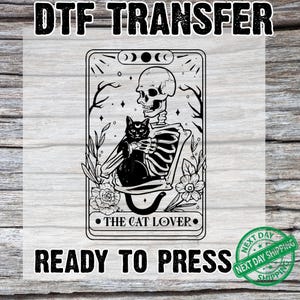 May include: Black and white DTF transfer featuring a skeleton holding a black cat, with the text "THE CAT LOVER." The design includes floral and moon phase elements. The words "DTF TRANSFER" and "READY TO PRESS" are also present.