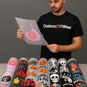 Puede incluir: Una persona sostiene una hoja impresa con un dise&ntilde;o de flor rosa, con varias hojas de transferencia enrolladas debajo. Los rollos presentan varios dise&ntilde;os, incluyendo flores, calabazas, esqueletos y texto. La persona lleva una camiseta negra con "Custom DTF Wear".