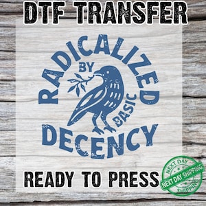 May include: A DTF transfer with the text "RADICALIZED BY BASIC DECENCY" in blue, surrounding a blue bird holding a sprig. The words "READY TO PRESS" are below the design. A green "NEXT DAY SHIPPING" stamp is in the lower right.