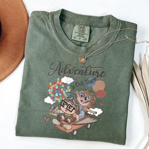 Puede incluir: Camiseta verde Comfort Colors con la frase "Adventure Is Out There" y un dibujo animado de dos personajes en un avi&oacute;n con globos. Un collar dorado est&aacute; drapeado sobre la camiseta.