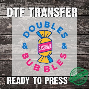 May include: DTF transfer design with the text "DOUBLES & BUBBLES" in pink and blue, surrounding a candy-shaped graphic with the word "BASEBALL" inside. The words "READY TO PRESS" are at the bottom.