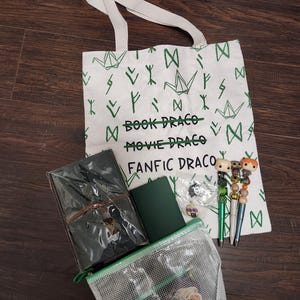 May include: A white canvas tote bag with green origami crane and rune patterns, featuring the text "BOOK DRACO", "MOVIE DRACO", and "FANFIC DRACO". Included are a dark green journal, a clear mesh zipper pouch, and three pens with character toppers.