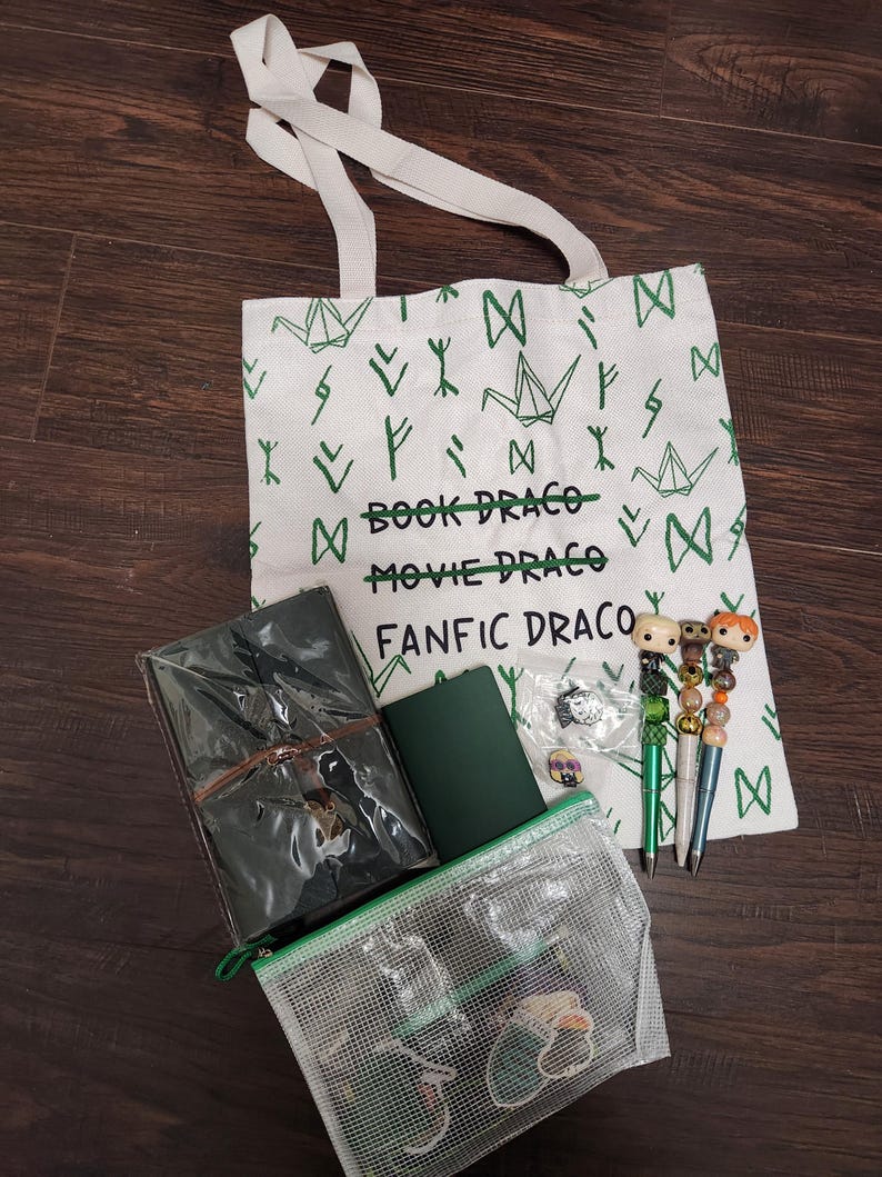May include: A white canvas tote bag with green origami crane and rune patterns, featuring the text "BOOK DRACO", "MOVIE DRACO", and "FANFIC DRACO". Included are a dark green journal, a clear mesh zipper pouch, and three pens with character toppers.