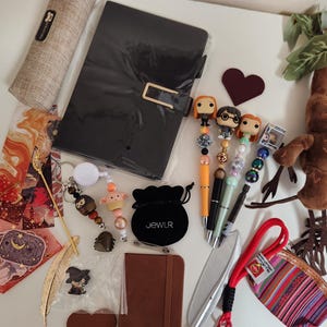 May include: A collection of items including a black notebook, a brown pencil case, four character-topped pens, a plush mandrake, a small black velvet pouch labeled JEWLR, a striped coin purse, a gold feather bookmark, and various small charms.