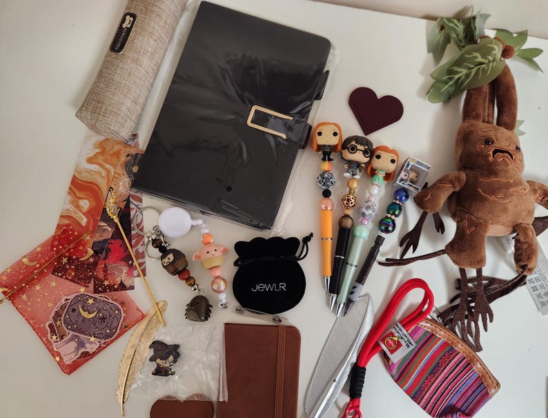 May include: A collection of items including a black notebook, a brown pencil case, four character-topped pens, a plush mandrake, a small black velvet pouch labeled JEWLR, a striped coin purse, a gold feather bookmark, and various small charms.