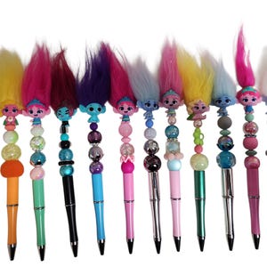 Puede incluir: Una colección de bolígrafos coloridos con muñecos Troll. Cada bolígrafo tiene un cuerpo de color diferente, un adorno de muñeco Troll y un diseño de cuentas. Los muñecos Troll tienen el pelo de diferentes colores, incluyendo rosa, amarillo y azul.