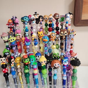 May include: A selection of character-topped pens displayed in a clear acrylic organiser. The pens feature various cartoon characters, with colourful beads and accents. The pens have black, blue, and green barrels. The pens are displayed in a clear acrylic organiser.