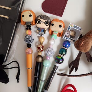May include: Four pens topped with miniature character figures and decorative beads. The pens feature orange, black, light green, and dark gray barrels. One pen includes a small Funko Pop box labeled Harry Potter 03 Hermione Granger.