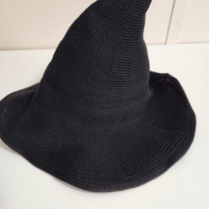 May include: A black knitted hat with a tall, pointed crown and a wide, slightly flared brim. The hat features a consistent, fine-textured knit pattern throughout, creating a structured yet flexible shape that tapers to a distinct point at the top.