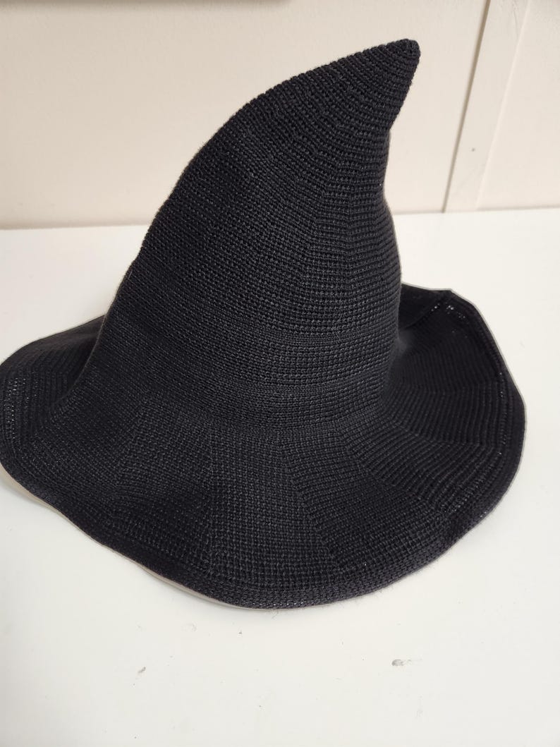May include: A black knitted hat with a tall, pointed crown and a wide, slightly flared brim. The hat features a consistent, fine-textured knit pattern throughout, creating a structured yet flexible shape that tapers to a distinct point at the top.