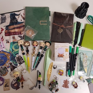 May include: A collection of stationery and accessories including a green notebook, a brown leather-style journal, a zippered pouch with wizarding illustrations, five character-themed pens, a miniature crystal ball box, stickers, bookmarks, and pins.