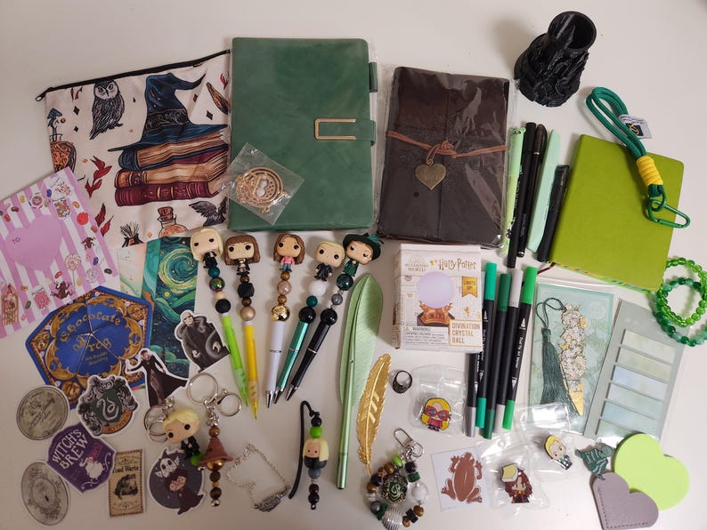 May include: A collection of stationery and accessories including a green notebook, a brown leather-style journal, a zippered pouch with wizarding illustrations, five character-themed pens, a miniature crystal ball box, stickers, bookmarks, and pins.