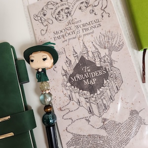 May include: A black pen with a decorative topper featuring a character figurine, with a green hat and dress. The pen is next to a map with the text "The Marauder's Map" and a green notebook. The words "Harry Potter" are visible.