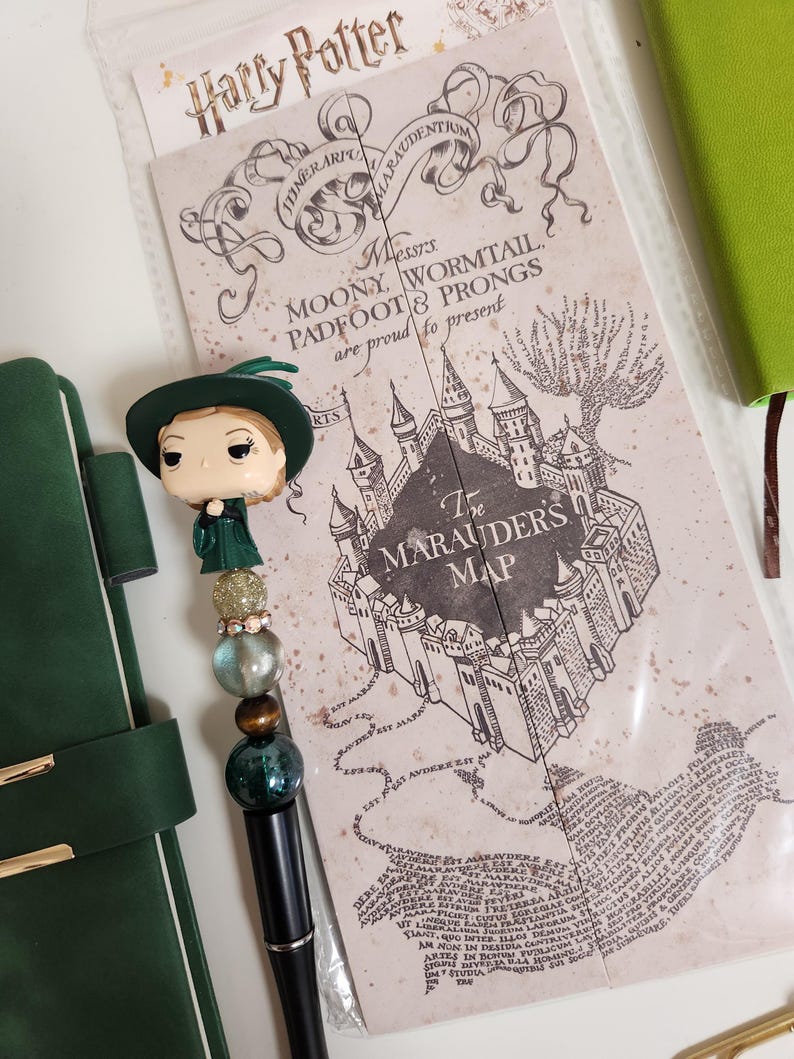 May include: A decorative pen topped with a miniature figurine of a character wearing a green hat and robe, accented by gold, clear, and dark green beads. Beside it lies a clear plastic sleeve containing a printed Harry Potter Marauder's Map.