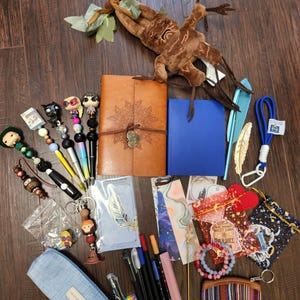 May include: A collection of stationery and accessories including a brown leather journal, a blue notebook, a plush mandrake, several beaded pens, bookmarks, a light blue pencil case, a small striped coin purse, and various charms and trinkets.