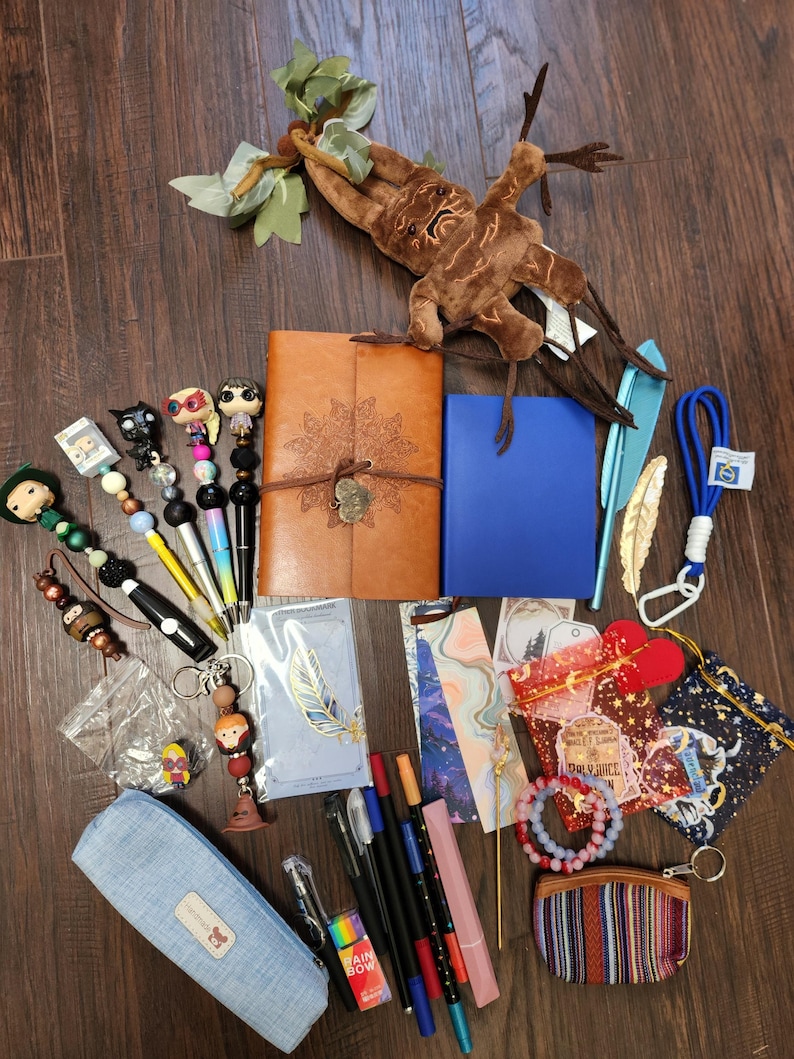 May include: A collection of stationery and accessories including a brown leather journal, a blue notebook, a plush mandrake, several beaded pens, bookmarks, a light blue pencil case, a small striped coin purse, and various charms and trinkets.