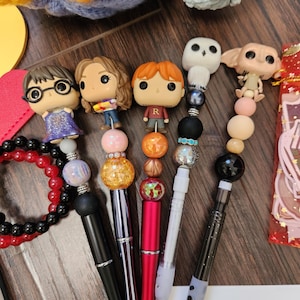 May include: Five pens featuring character toppers from the Harry Potter series. The pens include figures of Harry, Hermione, Ron, Hedwig, and Dobby. Each pen is decorated with various colorful beads, including glittery, metallic, and solid spheres.