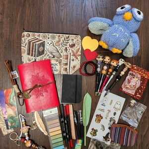 May include: A collection of stationery and accessories including a red leather journal, a zippered pouch with owl and book illustrations, a blue crocheted owl, decorative pens, bookmarks, stickers, a small black notebook, and various small trinkets.