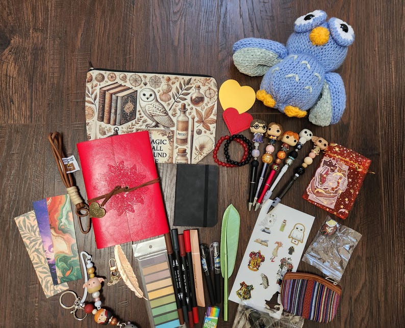 May include: A collection of stationery and accessories including a red leather journal, a zippered pouch with owl and book illustrations, a blue crocheted owl, decorative pens, bookmarks, stickers, a small black notebook, and various small trinkets.