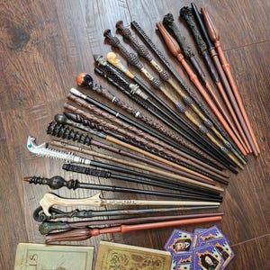 May include: A collection of fifteen toy wands in various brown, black, and silver tones, featuring diverse textures and handle designs. Two small rectangular books titled The Practical Pocket Book of Spells and The Practical Pocket Book of Herbology, plus several small hexagonal character cards are included.