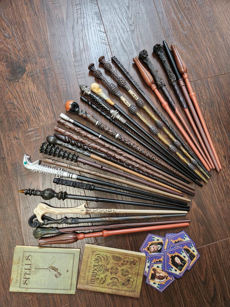 May include: A collection of fifteen toy wands in various brown, black, and silver tones, featuring diverse textures and handle designs. Two small rectangular books titled The Practical Pocket Book of Spells and The Practical Pocket Book of Herbology, plus several small hexagonal character cards are included.