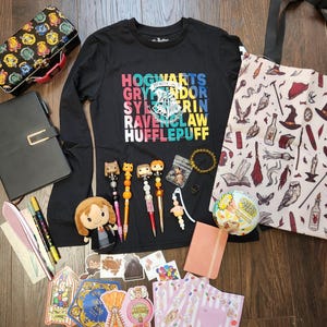 May include: A black long-sleeve shirt with a colorful Hogwarts graphic, a patterned tote bag, a lunch box, a black notebook, a plush doll, four decorative pens, a small pink journal, stickers, and various small accessories arranged on a dark surface.