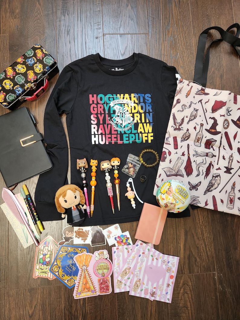 May include: A black long-sleeve shirt with a colorful Hogwarts graphic, a patterned tote bag, a lunch box, a black notebook, a plush doll, four decorative pens, a small pink journal, stickers, and various small accessories arranged on a dark surface.
