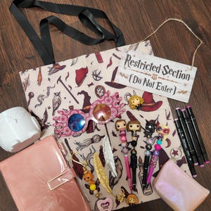 May include: A beige tote bag with a magical-themed print, a white owl mug, a pink glossy notebook, pink-rimmed glasses, a Restricted Section sign, four character-topped pens, a small iridescent pouch, three black markers, and two feather keychains.