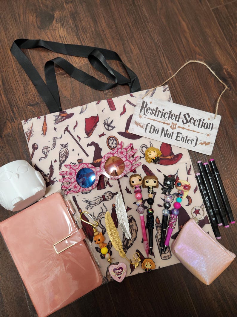 May include: A beige tote bag with a magical-themed print, a white owl mug, a pink glossy notebook, pink-rimmed glasses, a Restricted Section sign, four character-topped pens, a small iridescent pouch, three black markers, and two feather keychains.