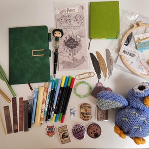 May include: A collection of stationery and craft items including a green journal, a Harry Potter Marauder's Map, a crochet blue owl, a wooden embroidery hoop, various pens, markers, feather-shaped bookmarks, stickers, and small decorative charms.
