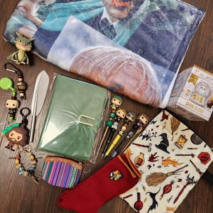 May include: A collection of merchandise including a printed fleece blanket, a green notebook, a snow globe box, a red sock with a crest, a patterned pouch, a small coin purse, four character pens, a metal letter opener, and several keychains.