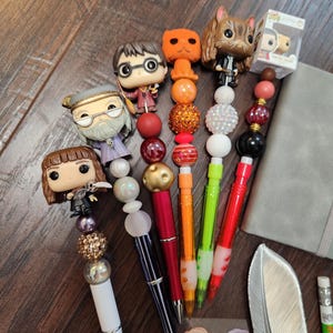 Peut inclure: Plusieurs stylos décoratifs ornés de figurines de personnages. Les stylos sont de différentes couleurs, dont le blanc, le rouge, l'orange et le vert, avec des perles décoratives. Une petite boîte Funko Pop est visible, ainsi qu'une plume et un carnet.