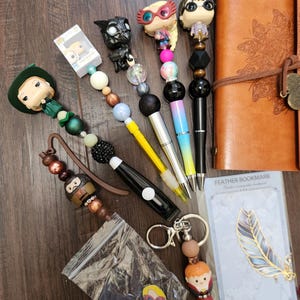 May include: A collection of character-themed pens and accessories including a brown leather journal, a feather bookmark in packaging, and several pens topped with miniature figures featuring colorful beads in various textures and finishes.