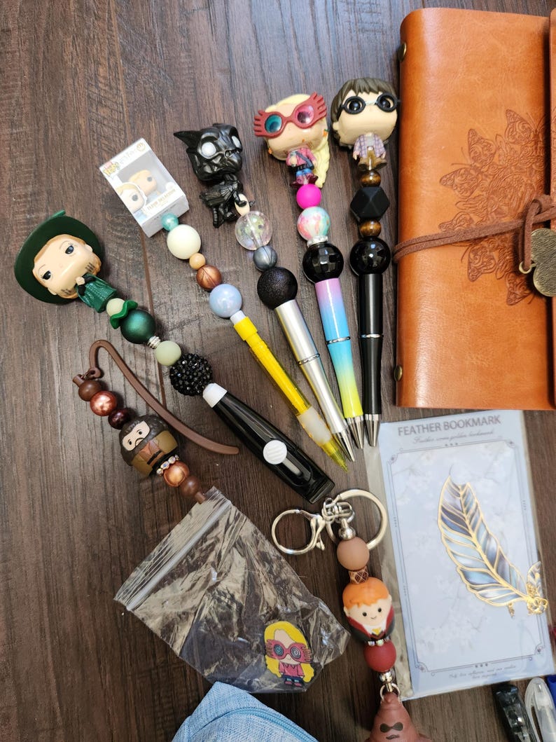 May include: A collection of character-themed pens and accessories including a brown leather journal, a feather bookmark in packaging, and several pens topped with miniature figures featuring colorful beads in various textures and finishes.