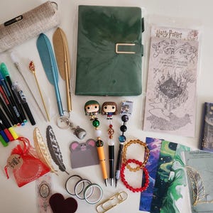 May include: A collection of stationery items including a green notebook, a beige pencil case, various colored markers, feather-shaped pens, beaded pens with character toppers, a Marauder's Map, bookmarks, a small notebook, and decorative accessories.