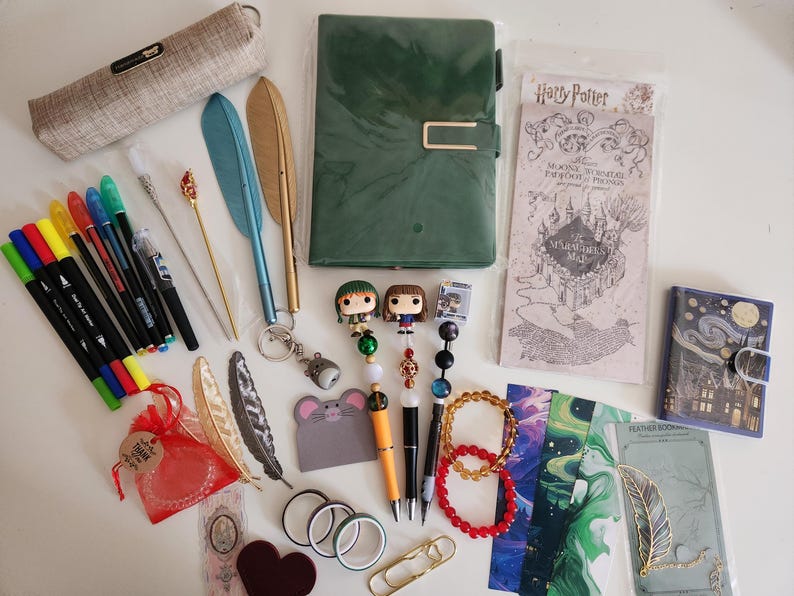 May include: A collection of stationery items including a green notebook, a beige pencil case, various colored markers, feather-shaped pens, beaded pens with character toppers, a Marauder's Map, bookmarks, a small notebook, and decorative accessories.