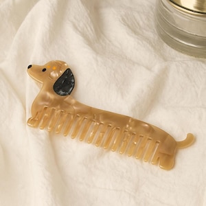 May include: A dachshund-shaped comb in shades of tan and brown, with black ear accents and a tail. The comb's teeth are evenly spaced, and the design is set against a white, textured background. A decorative item for hair care.