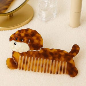 K&ouml;nnte beinhalten: Ein braun-beiger Kamm in Hundeform mit Schildpattmuster. Der Kamm hat ein wei&szlig;es Gesicht und liegt. Die Z&auml;hne des Kamms sind sichtbar. Der Kamm ist ein Sch&ouml;nheitsaccessoire.