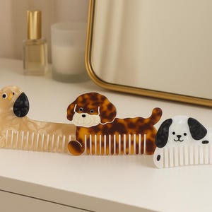 May include: Three decorative dog-shaped hair combs in a row. The first is a light tan color with black ears, the second is tortoiseshell, and the third is white with black spots and ears. The combs are on a white surface.