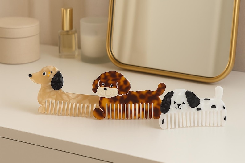 K&ouml;nnte beinhalten: Drei dekorative Haark&auml;mme in Hundeform in Creme, Schildpatt und Wei&szlig; mit schwarzen Akzenten. Die K&auml;mme befinden sich auf einer wei&szlig;en Oberfl&auml;che, mit einem Spiegel und anderen Objekten im Hintergrund.
