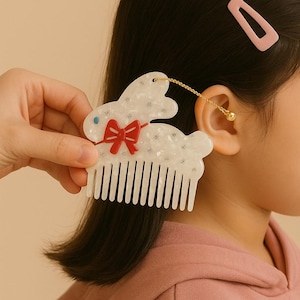 K&ouml;nnte beinhalten: Ein wei&szlig;er, hasenf&ouml;rmiger Haarkamm mit roter Schleife und goldener Kette. Der Kamm hat ein dekoratives Design mit kleinen Verzierungen. Eine rosa Haarspange ist im Hintergrund zu sehen. Der Kamm wird in der N&auml;he des Ohrs einer Person gehalten.