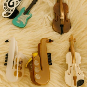 Puede incluir: Colección de broches con forma de instrumentos musicales. Incluye una clave de sol en blanco y negro, una guitarra turquesa, un violín marrón, un saxofón blanco, un saxofón marrón y un violín blanco. Los broches están sobre una superficie texturizada color crema.