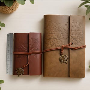 May include: Two brown leather journals with embossed maple leaf designs. The journals are secured with a brown cord and a metal leaf charm. One journal is smaller than the other. A ruler is next to the smaller journal.
