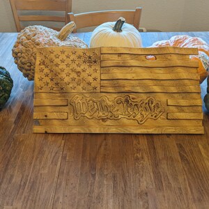 Puede incluir: Un letrero de madera con la bandera estadounidense con las palabras "We the People" talladas en la parte inferior. El letrero está rodeado de calabazas y calabacines.