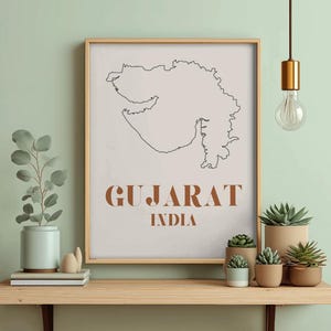 May include: A framed print featuring a minimalist outline of the Gujarat region of India, with the word "GUJARAT" and "INDIA" in a brown font. The print is displayed on a wooden shelf with potted plants and a hanging light fixture.