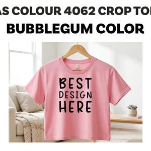 Puede incluir: Un top corto rosa chicle con el texto "BEST DESIGN HERE" en negro. La camiseta está colgada de una percha de madera. La parte superior es de cuello redondo y parece estar hecha de un material suave.