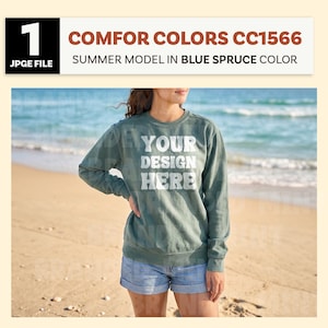 May include: A person wearing a blue spruce-colored sweatshirt and denim shorts stands on a sandy beach. The sweatshirt has the text "YOUR DESIGN HERE" in white. The image includes the text "COMFOR COLORS CC1566" and "SUMMER MODEL IN BLUE SPRUCE COLOR".