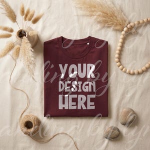 May include: A maroon t-shirt with the text "YOUR DESIGN HERE" in white, folded neatly on a cream-colored surface. Decorative elements include dried flowers in a vase, wooden beads, and twine.