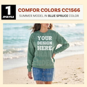 May include: A person wearing a green long-sleeve sweatshirt and a denim skirt stands on a beach. The sweatshirt has the text "YOUR DESIGN HERE" printed on the front. The image also includes the text "COMFOR COLORS CC1566 SUMMER MODEL IN BLUE SPRUCE COLOR".